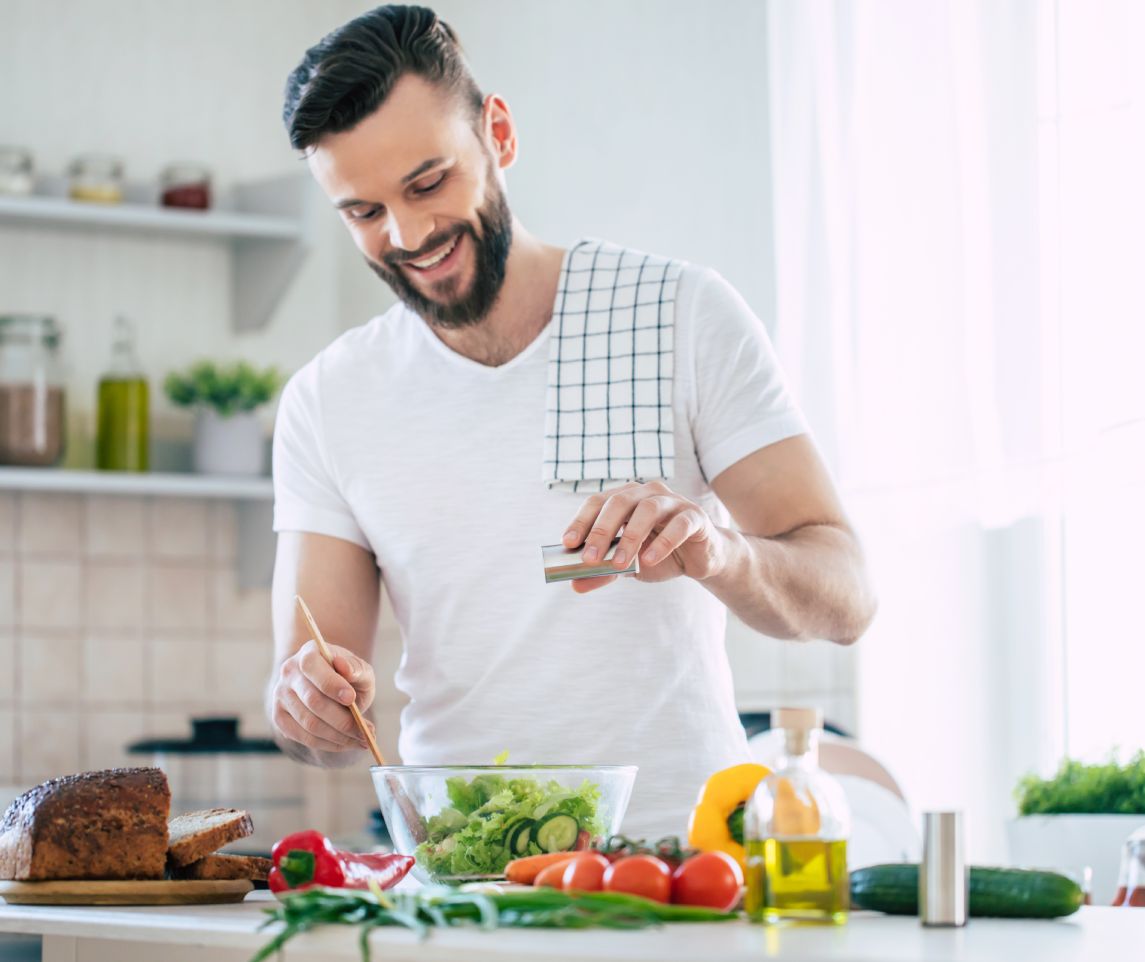 Handsome happy bearded man is preparing wonderful fresh vegan salad in the kitchen at home