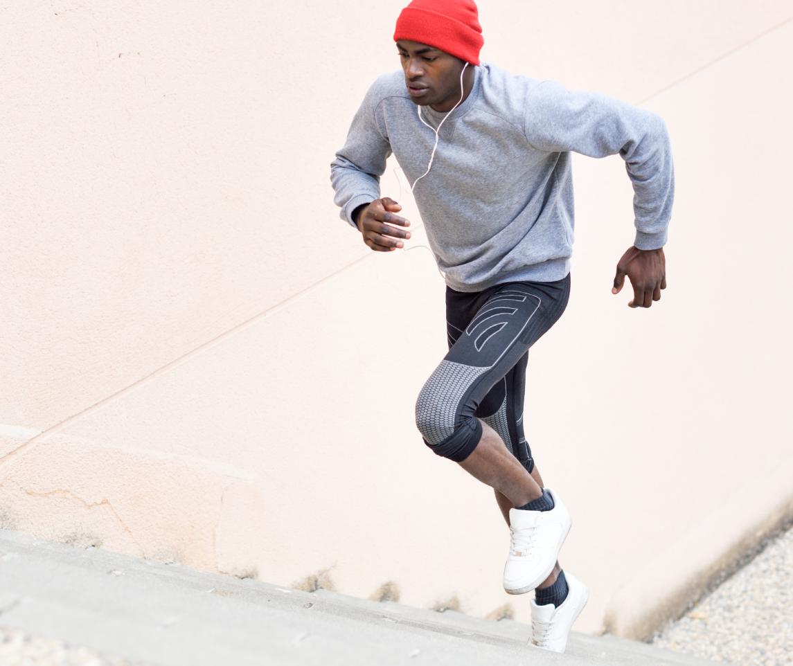 Black man running upstairs outdoors listening to music with white headphones. Young male exercising with city scape at the background.