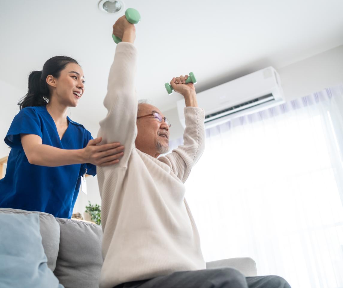 Asian older aged man doing physiotherapist with support from nurse. Senior elderly male sitting on sofa in living room using dumbbells workout exercise for patient with caregiver in nursing care.