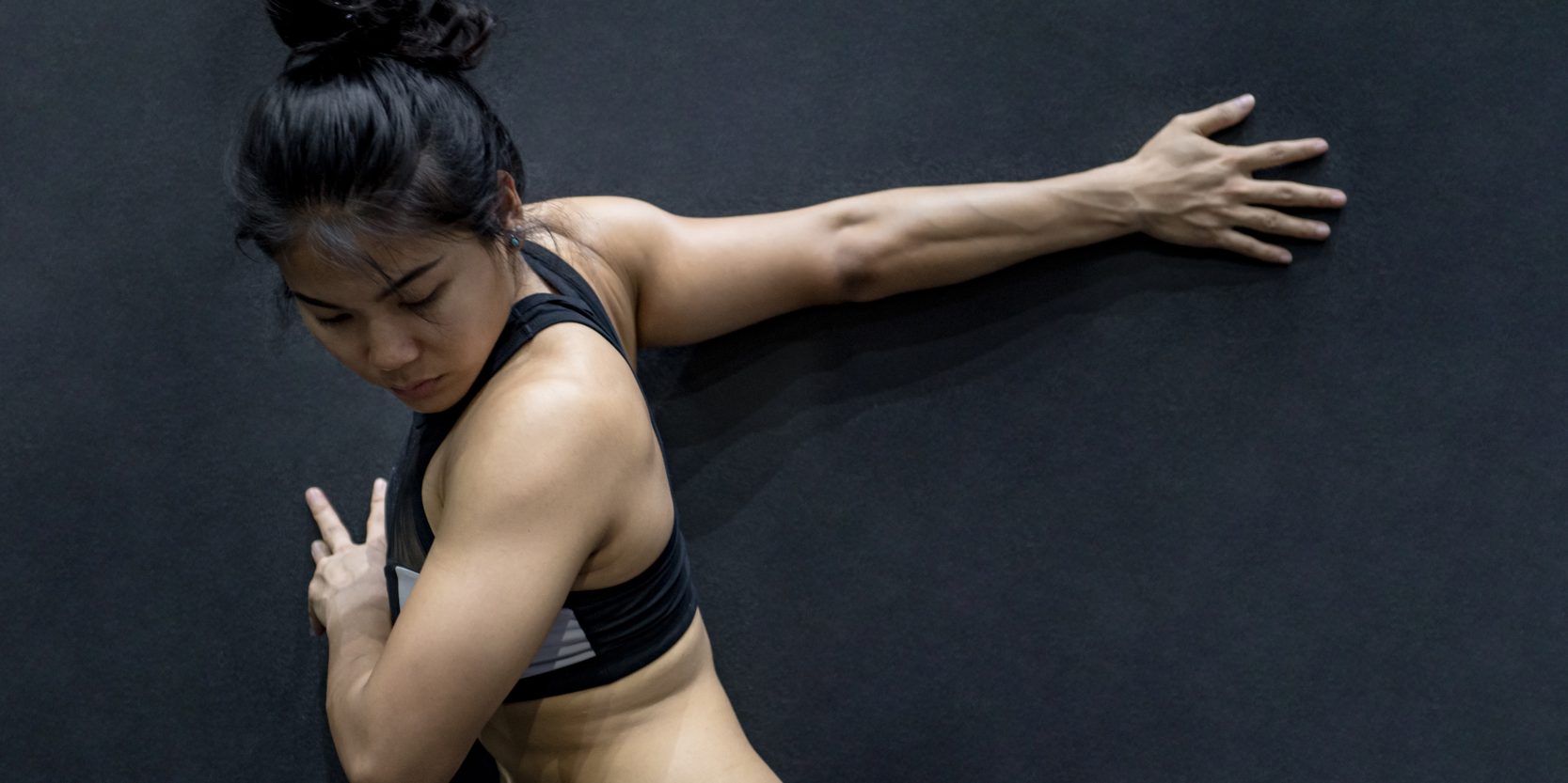 young asian woman wearing sportswear stretching her arm on black wall in fitness gym, exercise for healthy lifestyle concept