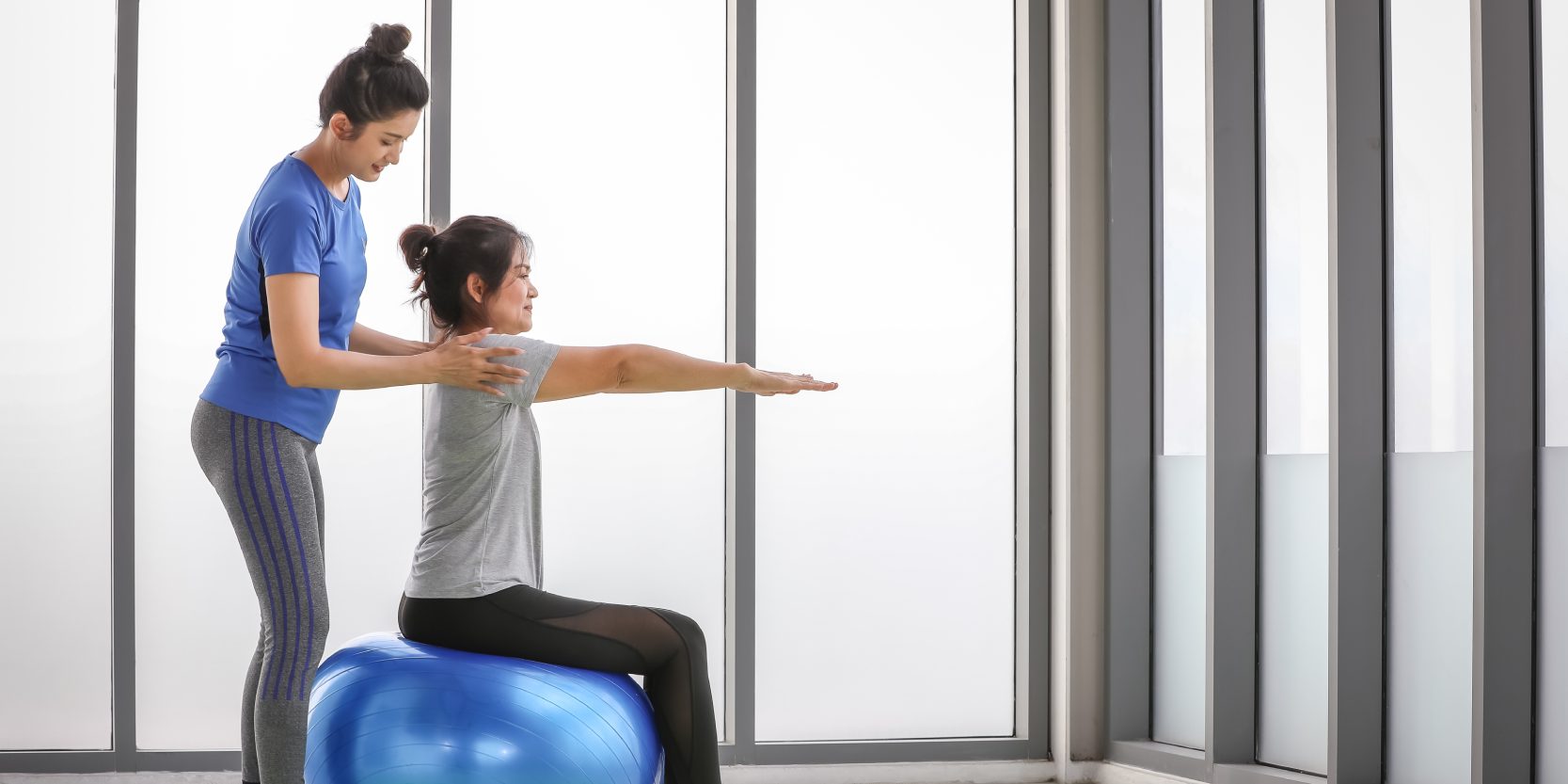 Beautiful exercise trainer enjoy helping Asian woman for yoga training by balancing body pose and shoulder position for arms stretching while sitting on fitness ball beside workout mat at sport gym.