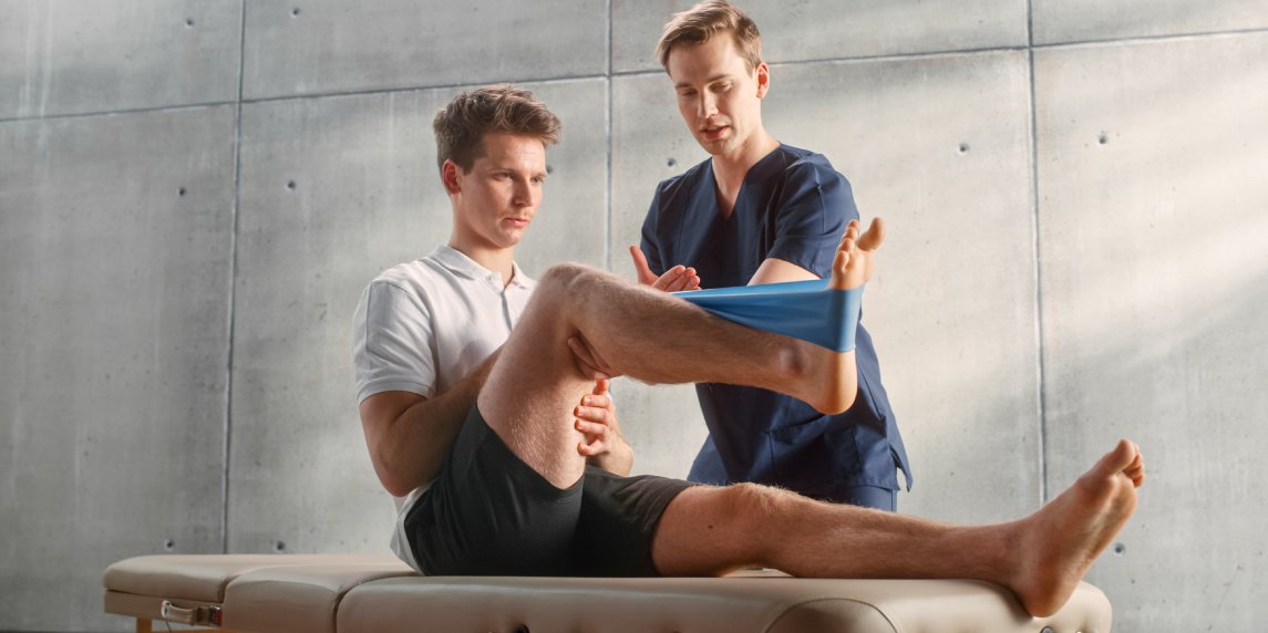 Sport Physiotherapy Specialist Showing How to Stretch a Rubber Band on Specific Muscle Groups or Joints to Young Male Athlete. Sportsman Recovering from Injury, Undergoing Rehabilitation.