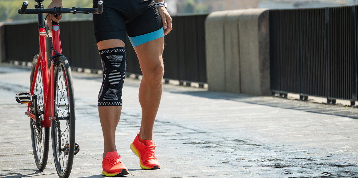 A person with knee braces poses beside a bike, representing solutions for cyclists dealing with chronic joint pain.