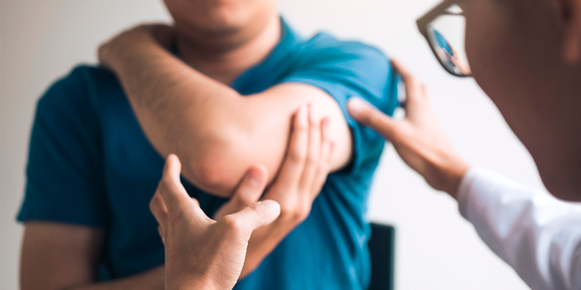 A man holds his arm in discomfort while another man observes him, likely discussing a sports injury.