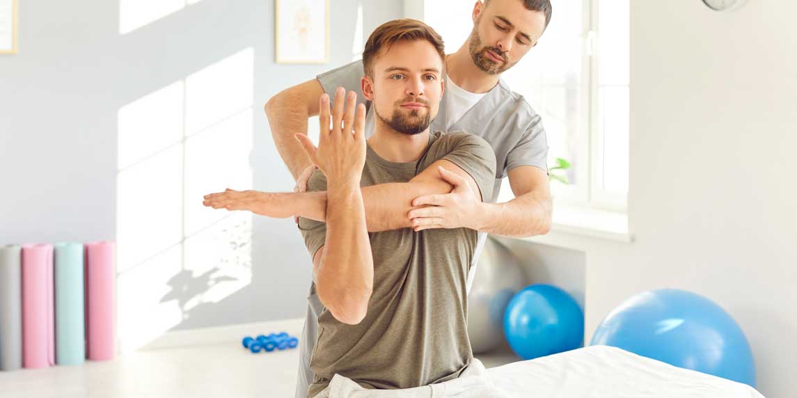 Male professional chiropractor or physical therapist working with Caucasian man's arm sitting on the couch in rehabilitation hospital. Physiotherapy and osteopathic medicine concept.